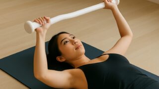 ヨガマットの上で仰向けになり、白いバーを胸の上で持ち上げている女性